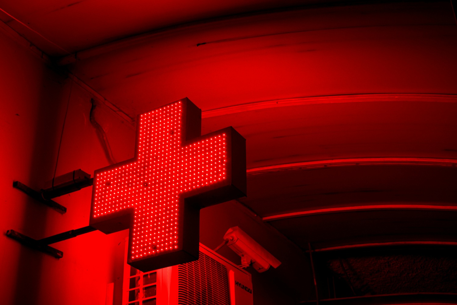 A close-up shot of a bright red neon pharmacy cross sign glowing against a dark background.