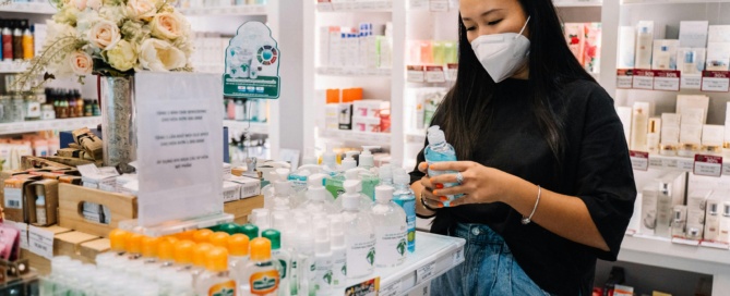 A female pharmacist wearing a blue face mask and sterile gloves carefully holds up a clear bottle inside a pharmacy.
