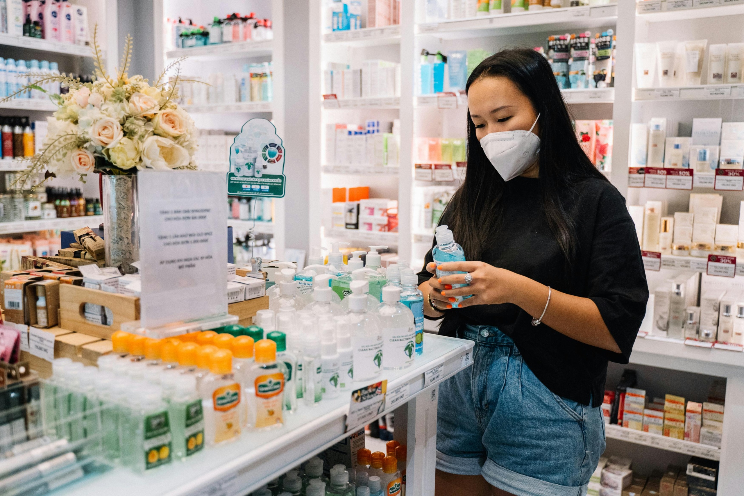 A female pharmacist wearing a blue face mask and sterile gloves carefully holds up a clear bottle inside a pharmacy.
