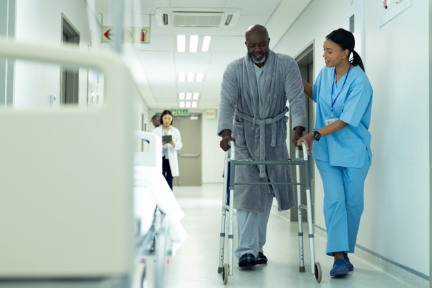 A nurse assists an elderly patient using a walker in a hospital hallway while medical staff continue working nearby, illustrating uninterrupted patient care in an active healthcare facility.